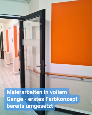 Das Bild zeigt eine halbgeöffnete Glastür mit schwarzem Rahmen, die einen Flur mit orange-weißer Farbgestaltung offenbart. Rechts neben der Tür ist eine große orangefarbene Fläche auf die Wand gemalt, oberhalb eines weißen Bereichs.  Ein Holzhandlauf verläuft an der Wand in niedriger Höhe. Über die Glastür zieht sich ein transparenter Streifen.  Am unteren Bildrand ist ein Texteinsatz sichtbar, der lautet: "Malerarbeiten in vollem Gange – erstes Farbkonzept bereits umgesetzt".