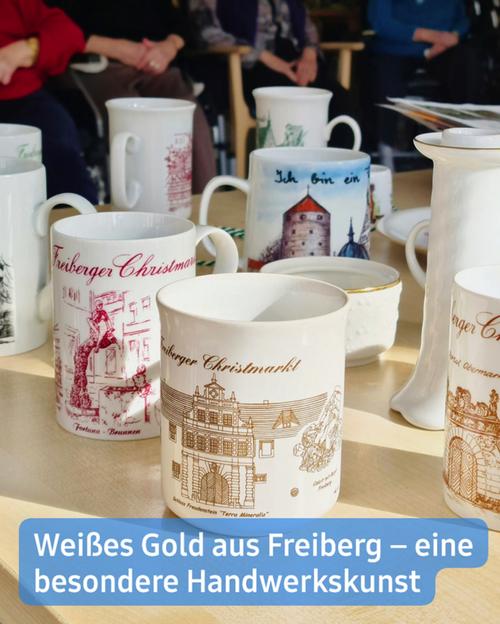 Eine hochwinkelige Nahaufnahme zeigt mehrere Tassen, die auf einer hellen Holzoberfläche stehen.  Die meisten Tassen sind weiß und mit goldenen Verzierungen sowie Schriftzügen versehen. Die Motive scheinen Gebäude und Szenen aus Freiberg, Deutschland, darzustellen. Sichtbarer Text auf den Tassen umfasst "Freiberger Christmarkt" und "Ich bin ein …".  Im Hintergrund sind, teilweise verschwommen, mehrere Personen in Stühlen zu sehen. Ebenso wie andere Objekte, darunter eine Vase und ein teilweise sichtbarer Tisch. Die insgesamt Atmosphäre vermittelt den Eindruck eines Marktes oder einer Messe.  Eine Beschriftung am unteren Rand lautet: "Weißes Gold aus Freiberg - eine besondere Handwerkskunst."