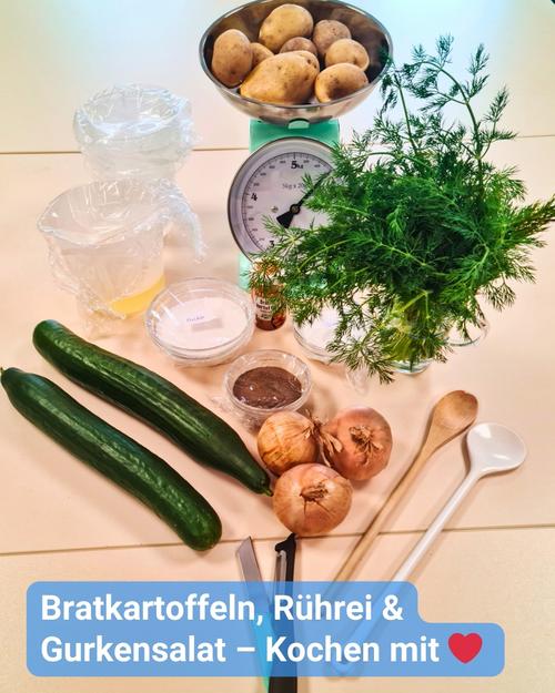 Nahaufnahme eines Tisches mit Lebensmitteln und Kochutensilien, der für das Kochen vorbereitet ist.  Im Mittelpunkt stehen verschiedene Zutaten, die ordentlich angeordnet sind: Kartoffeln auf einer Waage, ein Bund Dill, Gurken, Zwiebeln sowie vorbereitete Zutaten für einen Gurkensalat und Rührei in kleinen Behältern. Zusätzlich sind zwei transparente Kunststoffbehälter zu sehen, die möglicherweise Wasser oder andere Flüssigkeiten enthalten, sowie einige Küchenutensilien, darunter Holzlöffel und ein metallisches Utensil, das wie ein Buttermesser oder Spachtel aussieht. Der Hintergrund zeigt eine hellfarbige Tischoberfläche.  Eine Bildunterschrift am unteren Rand des Bildes, lautet: "Bratkartoffeln, Rührei & Gurkensalat – Kochen mit Herz"