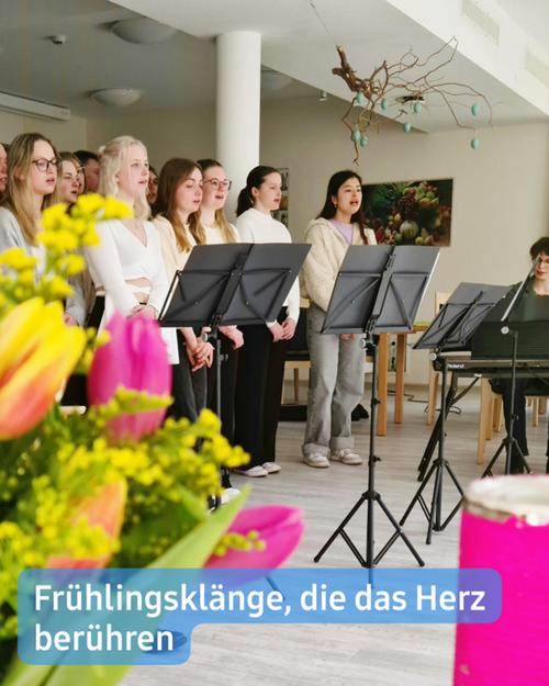Ein Chor junger Frauen, gekleidet in weiße und schwarze Oberteile, steht in einer Reihe und blickt dem Betrachter entgegen. Sie halten Notenblätter in den Händen und scheinen zu singen. Vor ihnen sind Notenständer aufgestellt. Der Raum ist hell und luftig, mit weißen Wänden und einer hohen Decke. Ein großes, astähnliches Kunstwerk hängt von der Decke.  Im Vordergrund befindet sich ein Strauß bunter Frühlingsblumen, der teilweise den unteren Teil des Chors verdeckt. Die Blumen zeigen überwiegend Gelb-, Pink- und Orangetöne.  Eine Überschrift am unteren Rand lautet: "Frühlingsklänge, die das Herz berühren"
